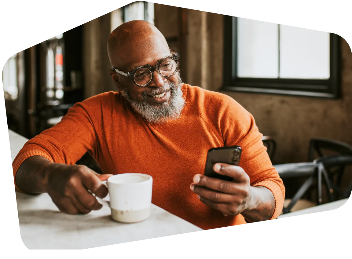 A bald man with glasses smiling at his phone while holding a coffee cup in a café.