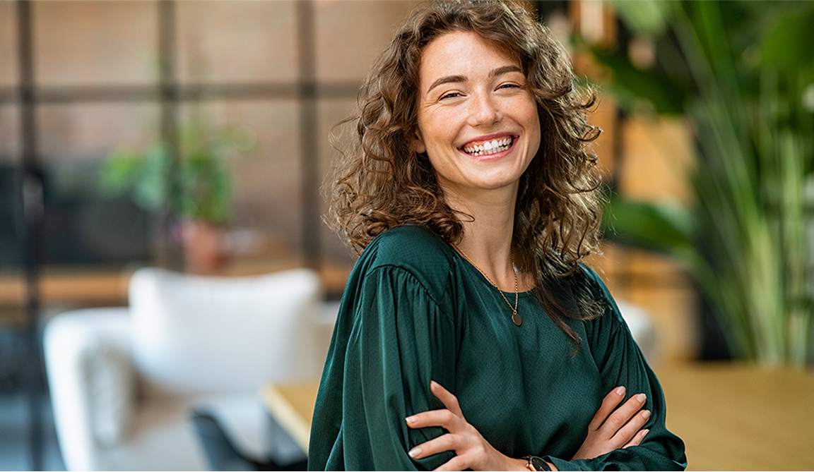 A cheerful young woman with curly hair wearing a green blouse, standing with arms crossed and smiling.