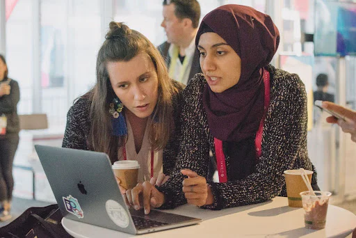 Collaborating on a Project at a Coffee Table Two women working together on a laptop at a table, one holding a coffee cup, in a public indoor space.