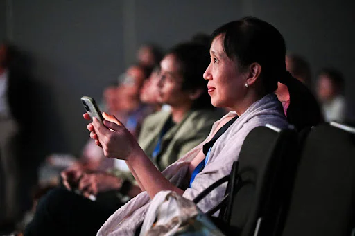 Attendees Engaged During a Conference Woman holding a phone, sitting among other attendees during a conference.
