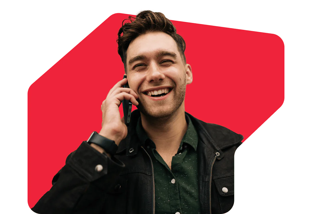 A smiling man holding a phone to his ear, wearing a dark jacket and green shirt with a red background.